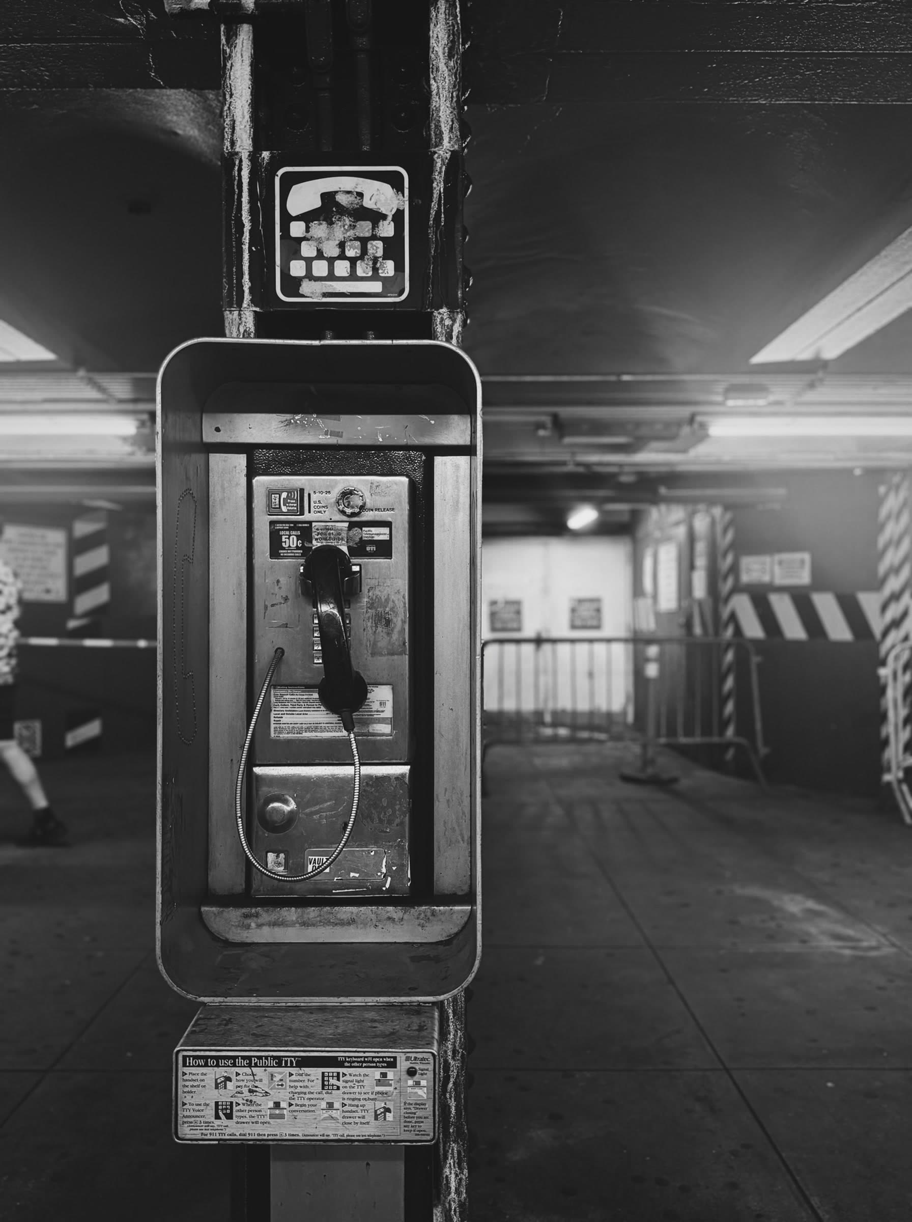 The payphone on its column in a subway station; someone passes at the edge of the frame.