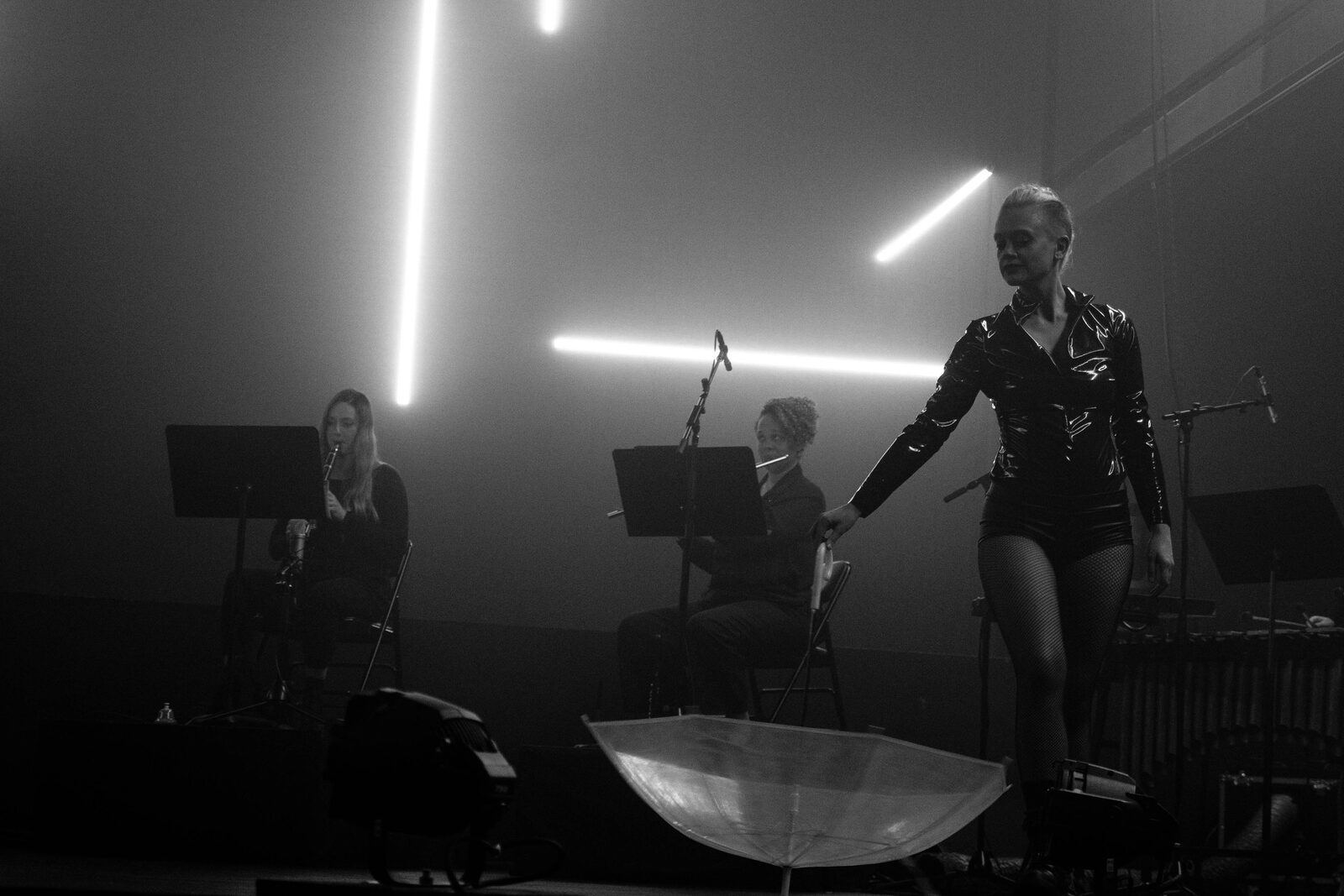 Performer in a clear umbrella under horizontal neon bars, flutist and musicians on stage in haze