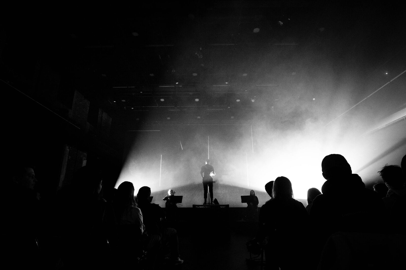 Silhouetted figure on a low stage, backlit by haze and a shaft of light above a seated audience