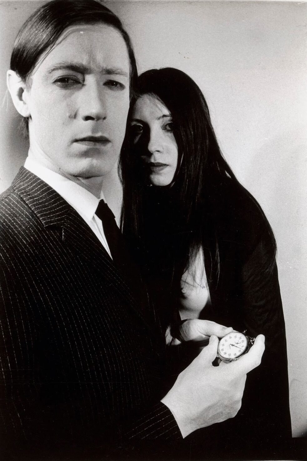 Black-and-white portrait still, man in pinstripe suit with woman behind holding a pocket watch