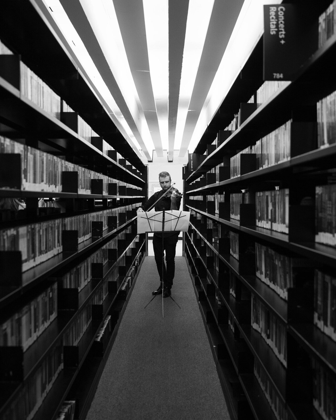 Whitney George with violin in a library aisle, black-and-white