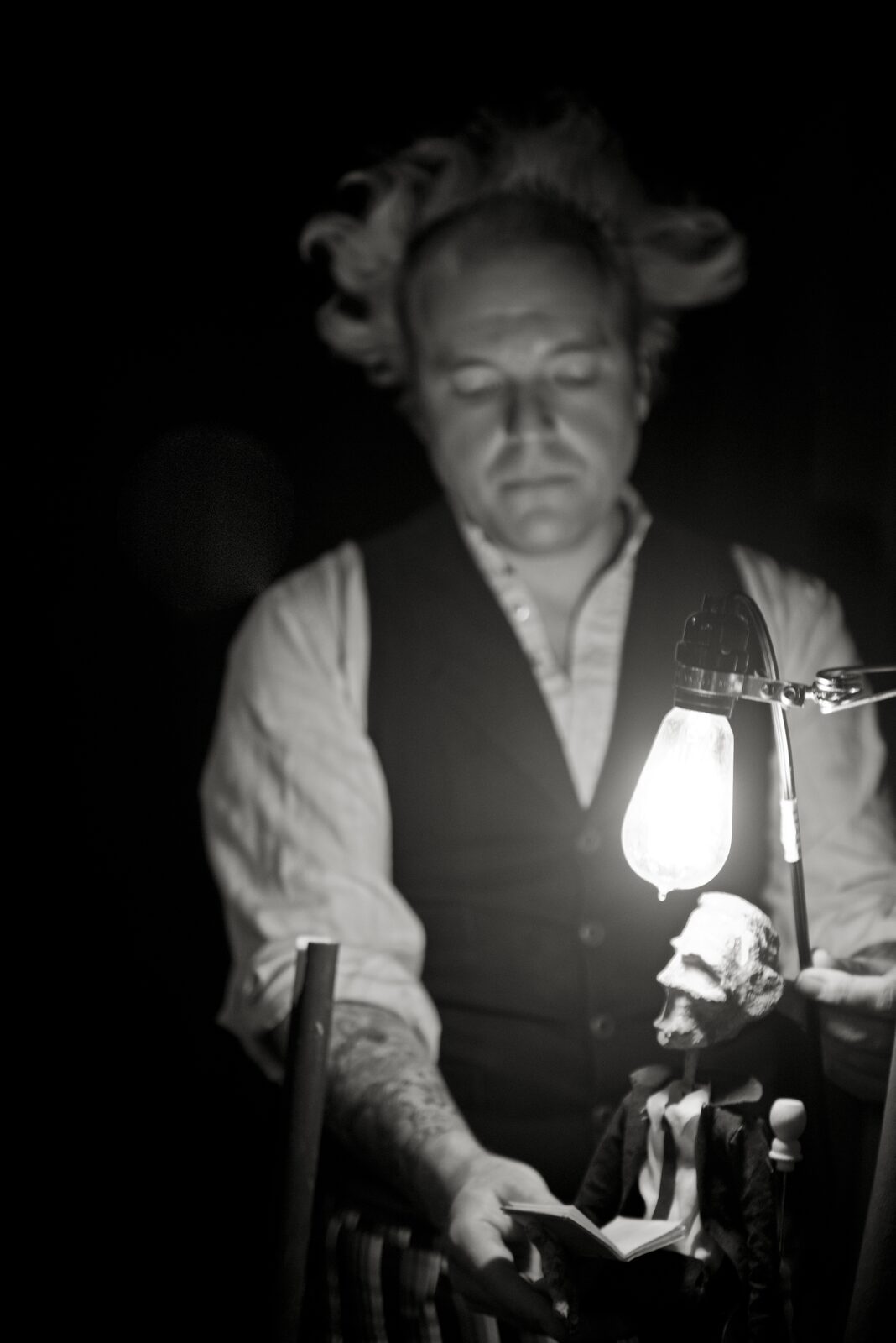 Puppeteer in waistcoat holding a small skull-like puppet under a lit Edison bulb, darkened stage